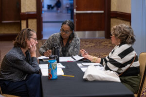 Three people engage in discussion and note-taking at a table in a conference setting.