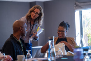 A woman speaks to a man while another woman smiles, unpacking a bag at a meeting table.