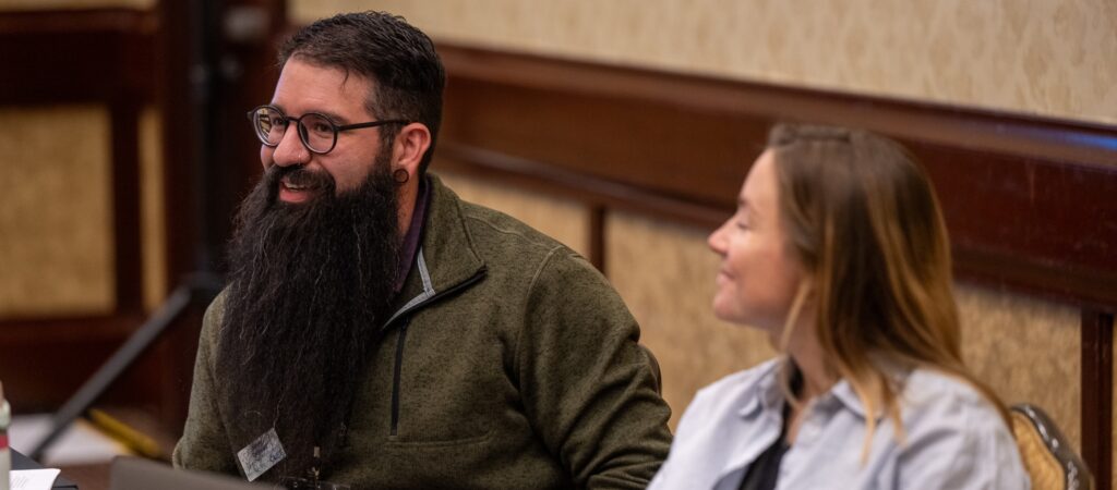 Two people sit at a table, engaged in conversation. One has a long beard and glasses, the other smiles.
