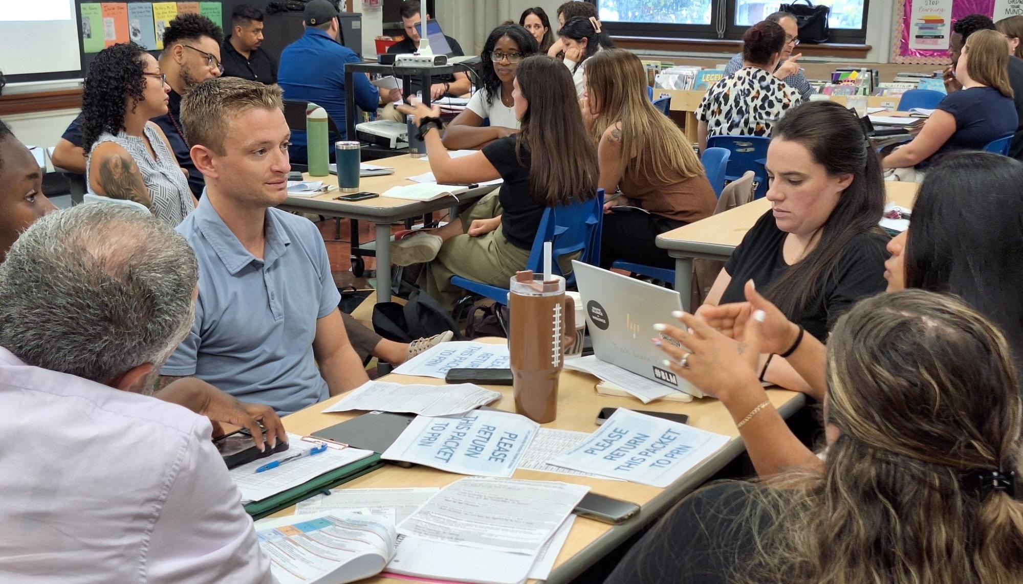 A group of people engages in discussion at tables, with papers and laptops scattered around.
