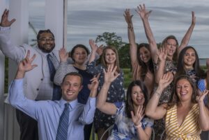 A group of ten people joyfully raise their hands, smiling and celebrating together outdoors.
