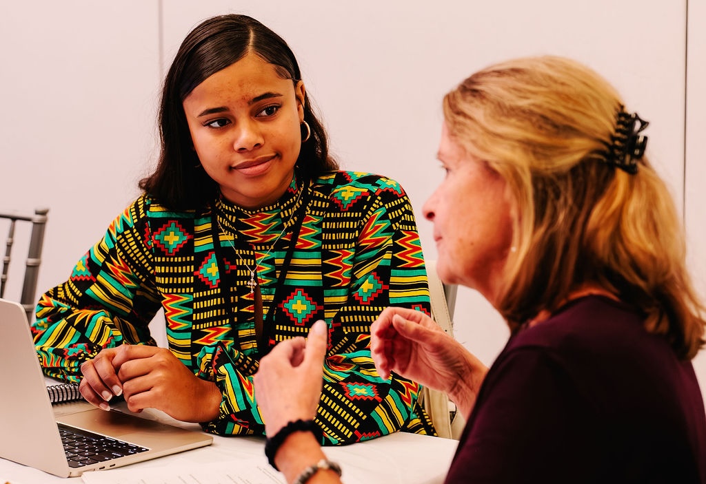 A young woman listens attentively to an older woman speaking, with a laptop on the table between them.