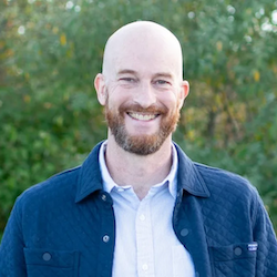 A smiling man with a beard stands outdoors, wearing a blue jacket over a white shirt, surrounded by greenery.