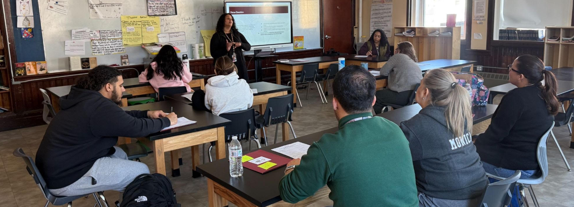 A teacher presents to a group of seated adults in a classroom setting.