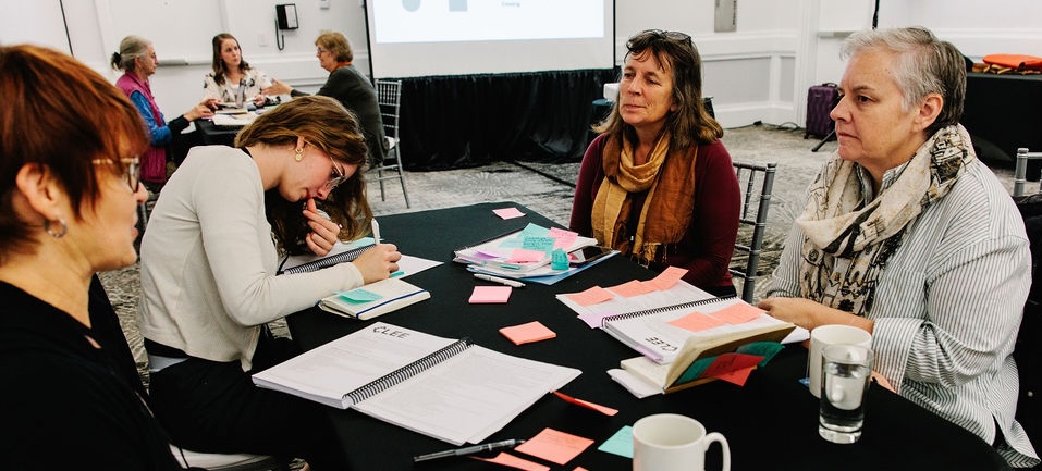 Four women engage in discussion at a table covered with notes and materials, with a presentation in the background.