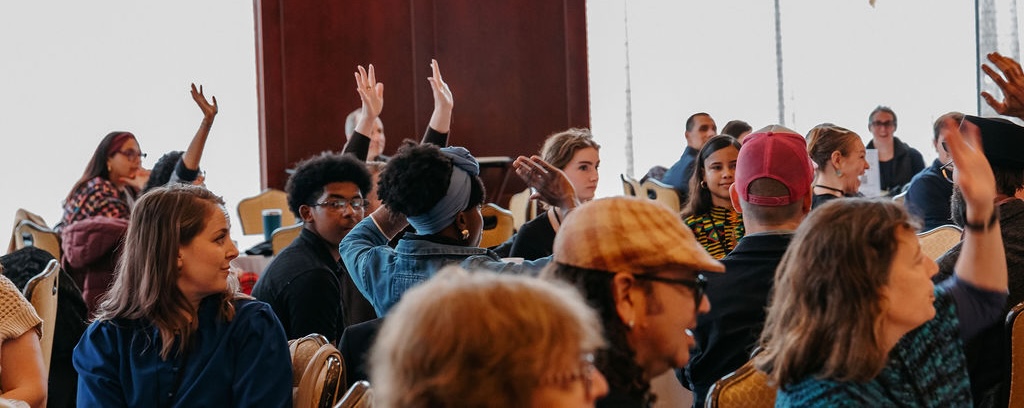 A group of educators holding their hands up in a large room at Fall meeting